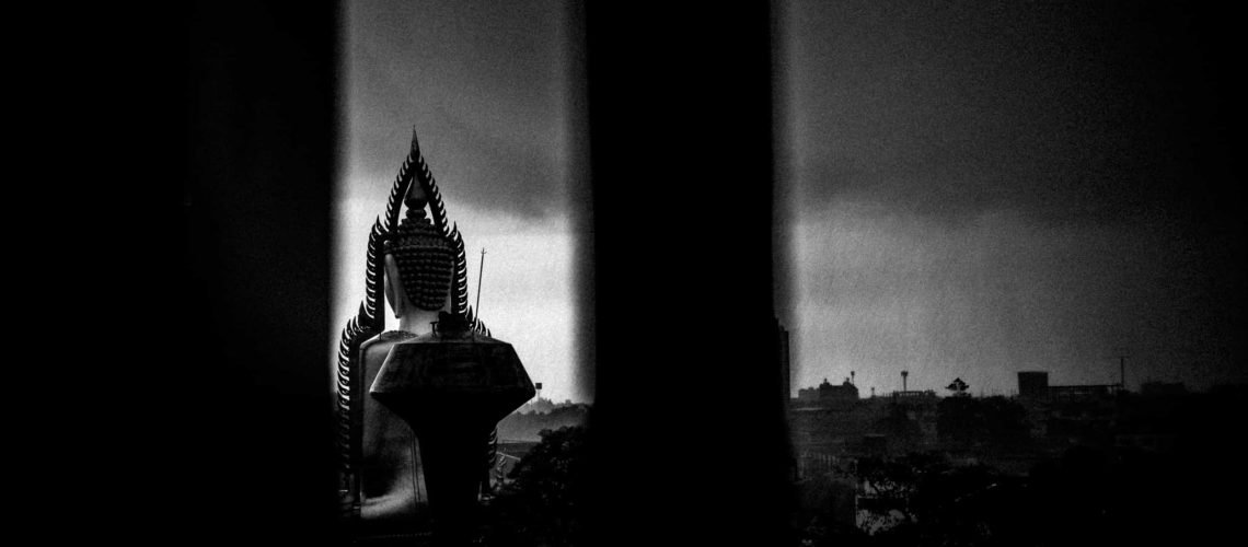 Silhouette of a large seated Buddha in Bangkok, Thailand, set against the darkness of an afternoon thunderstorm